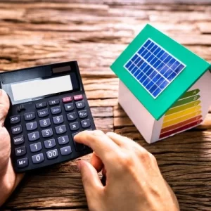 Homeowner using a calculator, calculating the cost of solar energy for his home. Model of a home with solar panel next to him. 